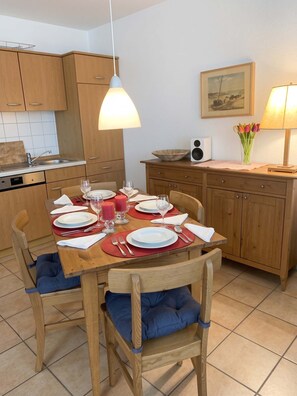 Een eethoek met een houten tafel voor vier personen, stoelen met blauwe kussens, een keuken op de achtergrond en een speaker bevestigd aan de muur.