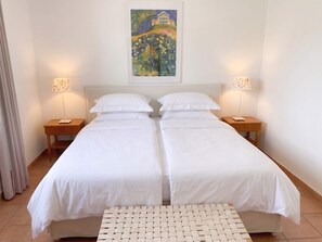 A double bed with white linens, two bedside tables with lamps, a woven bench, and a painting on the wall. - FeWo Frische Brise - Lauterbach (Putbus)