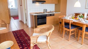 A compact kitchen with modern appliances, a dining area with wicker chairs, and a red patterned rug. - FeWo Frische Brise - Lauterbach (Putbus)