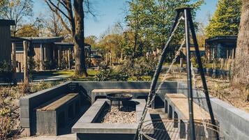 Een terras met beton, een vuurkorf, omgeven door bomen en een gebouw op de achtergrond.