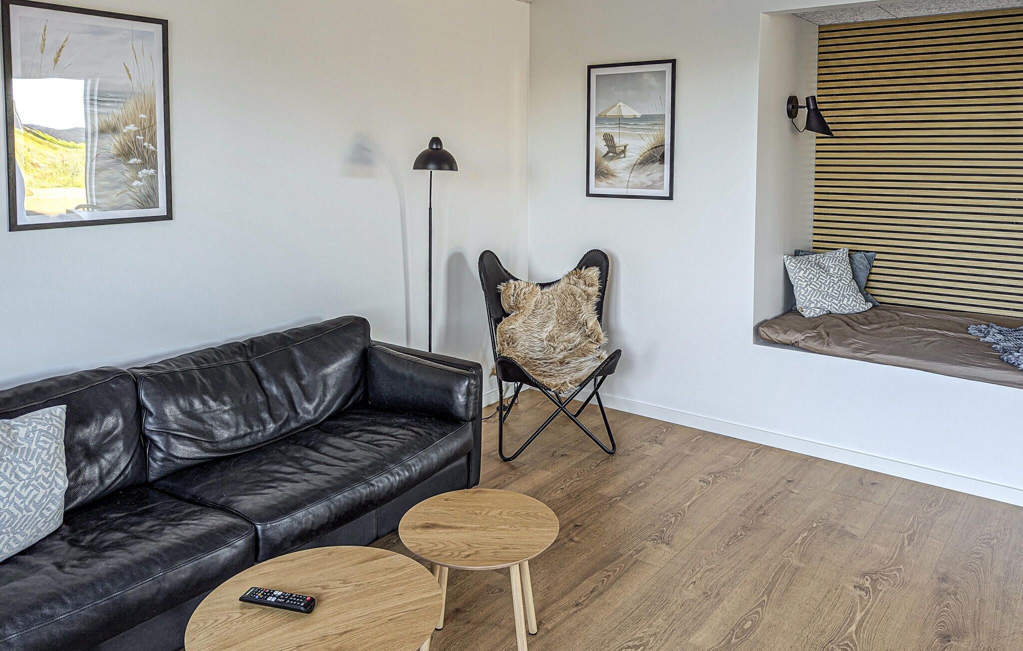 Una sala de estar con un sofá de cuero negro, dos mesitas de madera, un sillón cubierto de piel y un ventanal con persianas.