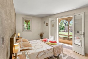 A bedroom with a bed, a nightstand, a lamp, a window, and an open door leading to a balcony. - Garden-Level Villa & Independent Guest House 'Strelitzia' Near the Beach (Sainte-Maxime)