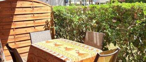 Een terras buiten met een tafel en stoelen, omgeven door groen en een wit gebouw op de achtergrond.