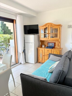 Ein Zimmer mit einer Couch, einem Fernseher auf einem Schrank, einem Kühlschrank, einer Holzvitrine und einer Glastür zu einem Außenbereich mit einem Fahrrad.