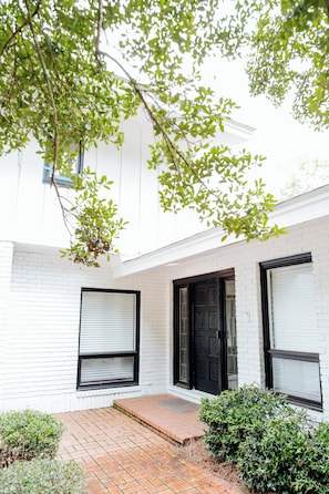 A white brick building with a black door and windows, a brick pathway, and green foliage.