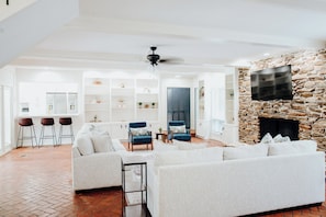 A spacious living room with a stone fireplace, a flat-screen TV, and a kitchen area in the background.