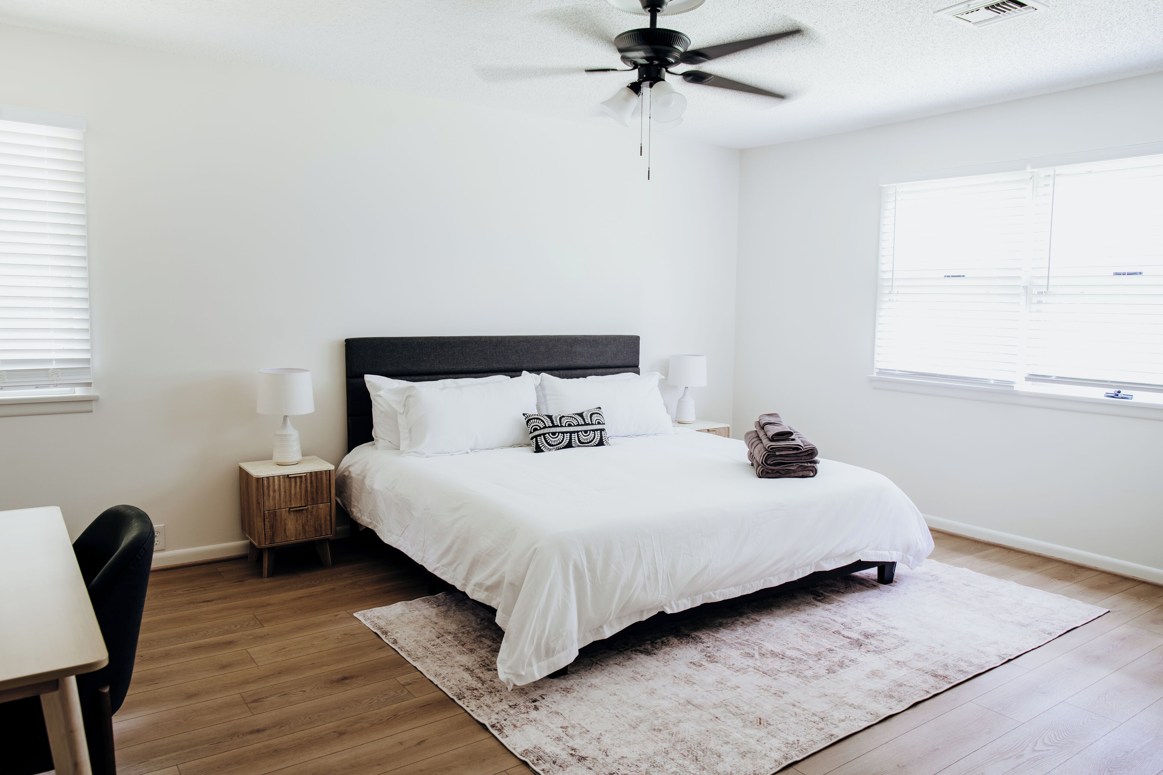 A bedroom with a bed, bedside lamps, a ceiling fan, a window, and a desk.