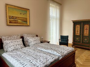 Ein Schlafzimmer mit einem Bett, einem Sessel, einem Nachttisch, einer Lampe, einem Gemälde und einem Schrank.
