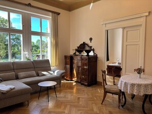 Ein Wohnzimmer mit einer Couch, einem kleinen Tisch, einem Holzschrank und einem Esstisch mit einer Blumen Tischdecke.