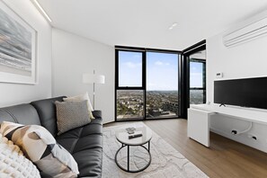 A modern living room with a black leather sofa, a round coffee table, a large flat-screen TV, and a city view through floor-to-ceiling windows. - City-view 1-bed Retreat Near Queen Vic (Melbourne)