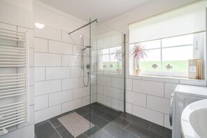 Uma casa de banho moderna com azulejos brancos, cabine de duche em vidro, máquina de lavar embutida e janela com vista para vegetação.