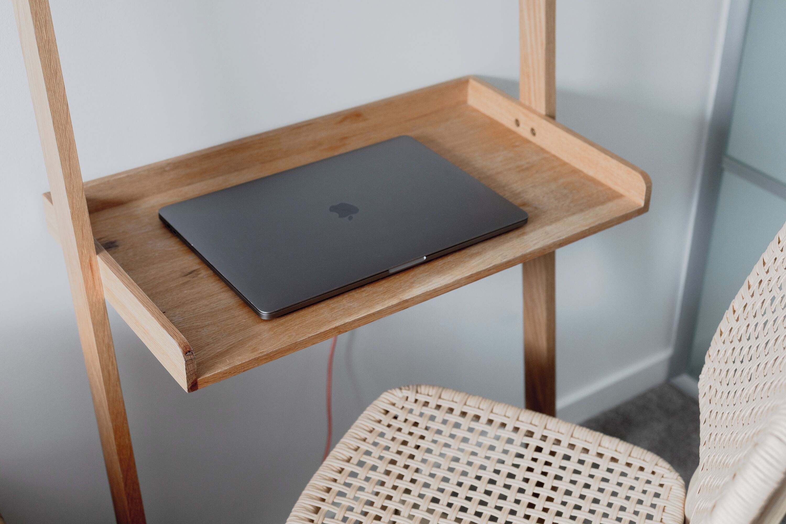 Un portátil sobre una estantería de madera, una silla de mimbre y una esquina de habitación con pared blanca.