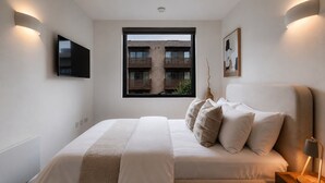 Een slaapkamer met een bed, een televisie, een raam met uitzicht op een gebouw en een nachtkastje met een lamp.