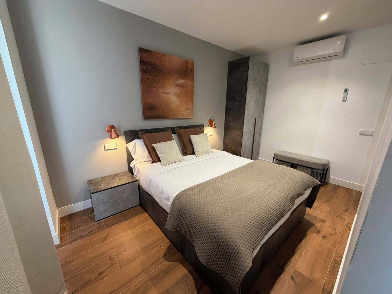 Un dormitorio moderno con una cama grande, lámparas de noche, aire acondicionado de pared y una banca.