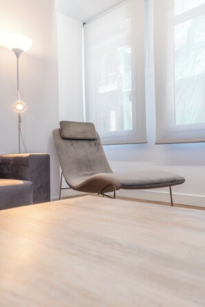 Um quarto moderno com uma poltrona cinza, um abajur e grandes janelas.