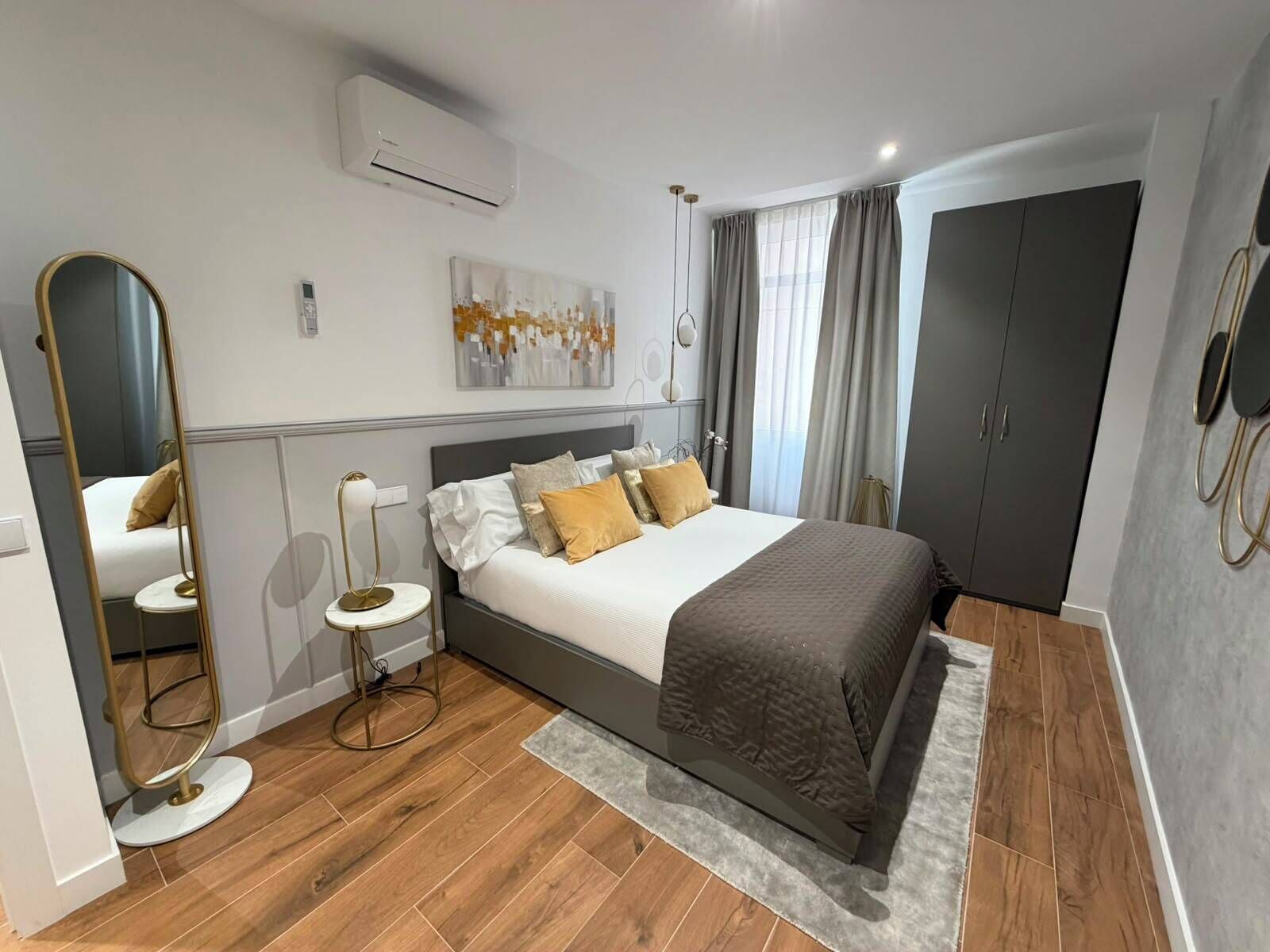 A modern bedroom with a bed, a round mirror, a bedside table, a wall-mounted air conditioner, and a wardrobe.