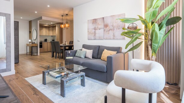 A modern living room with a sofa, a glass coffee table, and a chair. There is a dining area in the background with a table and chairs. - Apartamentos Príncipe de Canalejas (Ma)