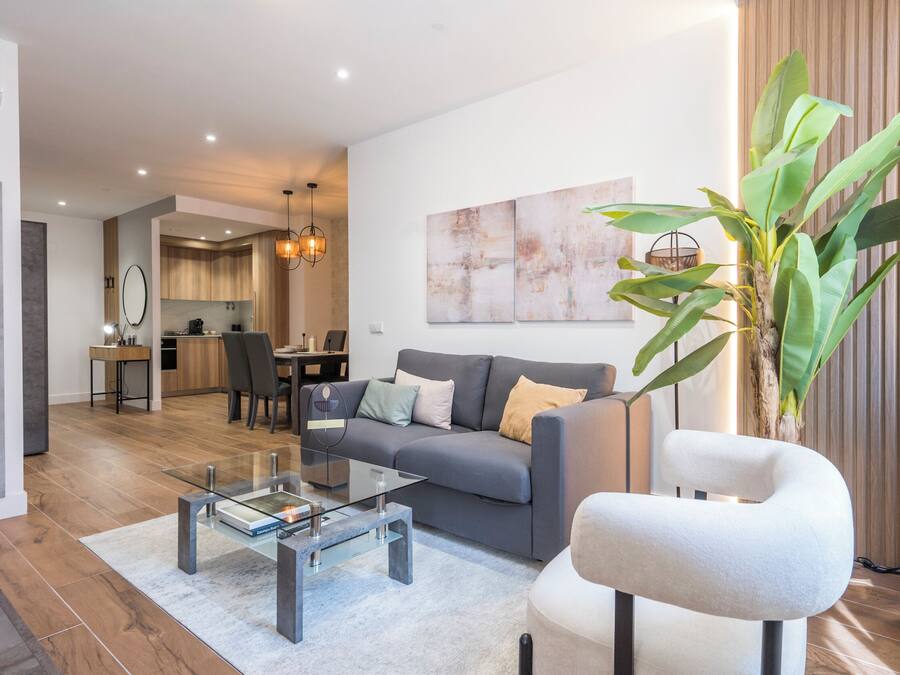 Una sala moderna con un sofá, una mesa de centro de vidrio y una silla. En el fondo, hay un comedor con una mesa y sillas.