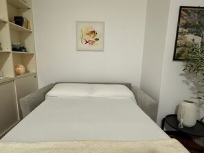 Una cama con ropa de cama blanca, una estantería con libros y una vela, y un cuadro en la pared.