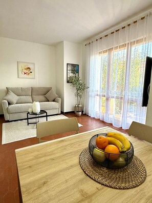 A living room with a sofa, a dining table with a fruit bowl, a potted plant, and a window with curtains.