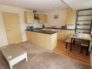 A compact kitchen with wooden cabinets, a microwave, a kettle, and a dining area with a table and chairs. - Twyni Bach (Burry Port)