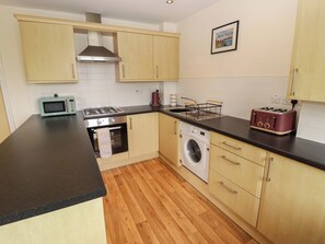 A kitchen with wooden cabinets, a microwave, a stove, a washing machine, and a toaster. - Twyni Bach (Burry Port)