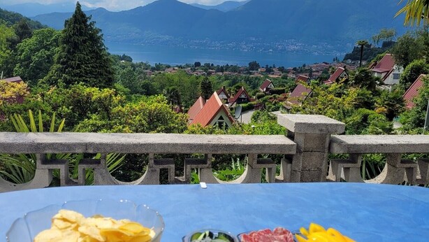 A table with snacks, cheese, and wine glasses set against a scenic view of mountains and a lake. - Casa Ines with Lake Maggiore View (Villaggio Olandese)