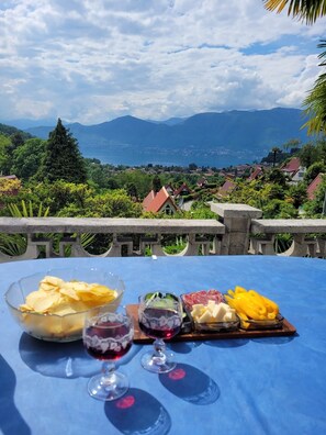A table with snacks, cheese, and wine glasses set against a scenic view of mountains and a lake. - Casa Ines with Lake Maggiore View (Villaggio Olandese)