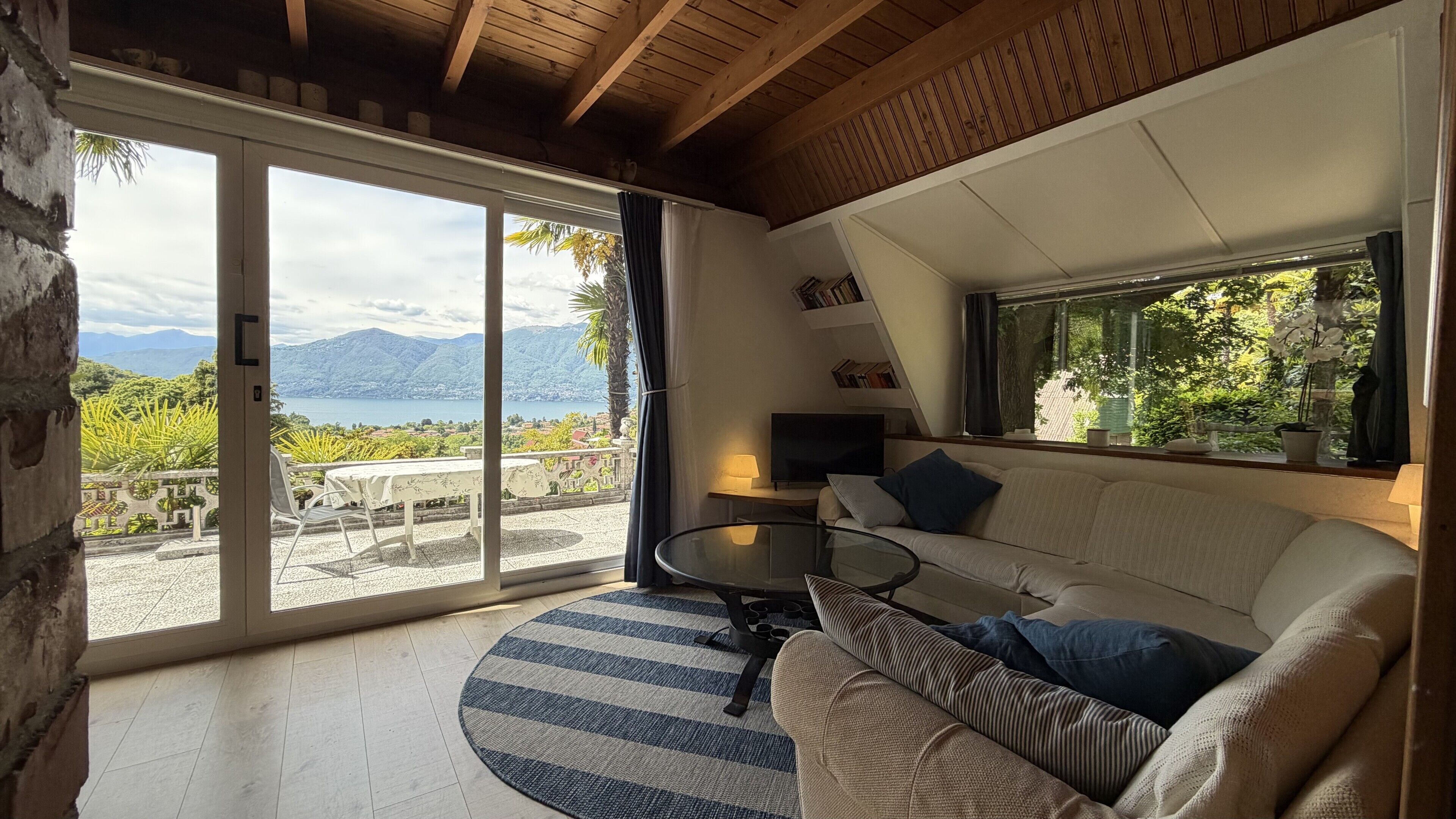 A modern living room with a sofa, a glass coffee table, a TV, and a view of mountains and a lake.