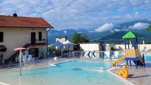A swimming pool with a children's play area, lounge chairs, and a building with a red-tiled roof. - Casa Ines with Lake Maggiore View (Villaggio Olandese)