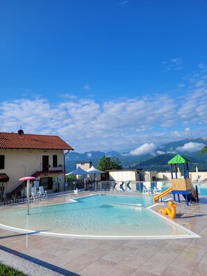 Una piscina con area giochi per bambini, lettini e un edificio con tetto in tegole rosse.