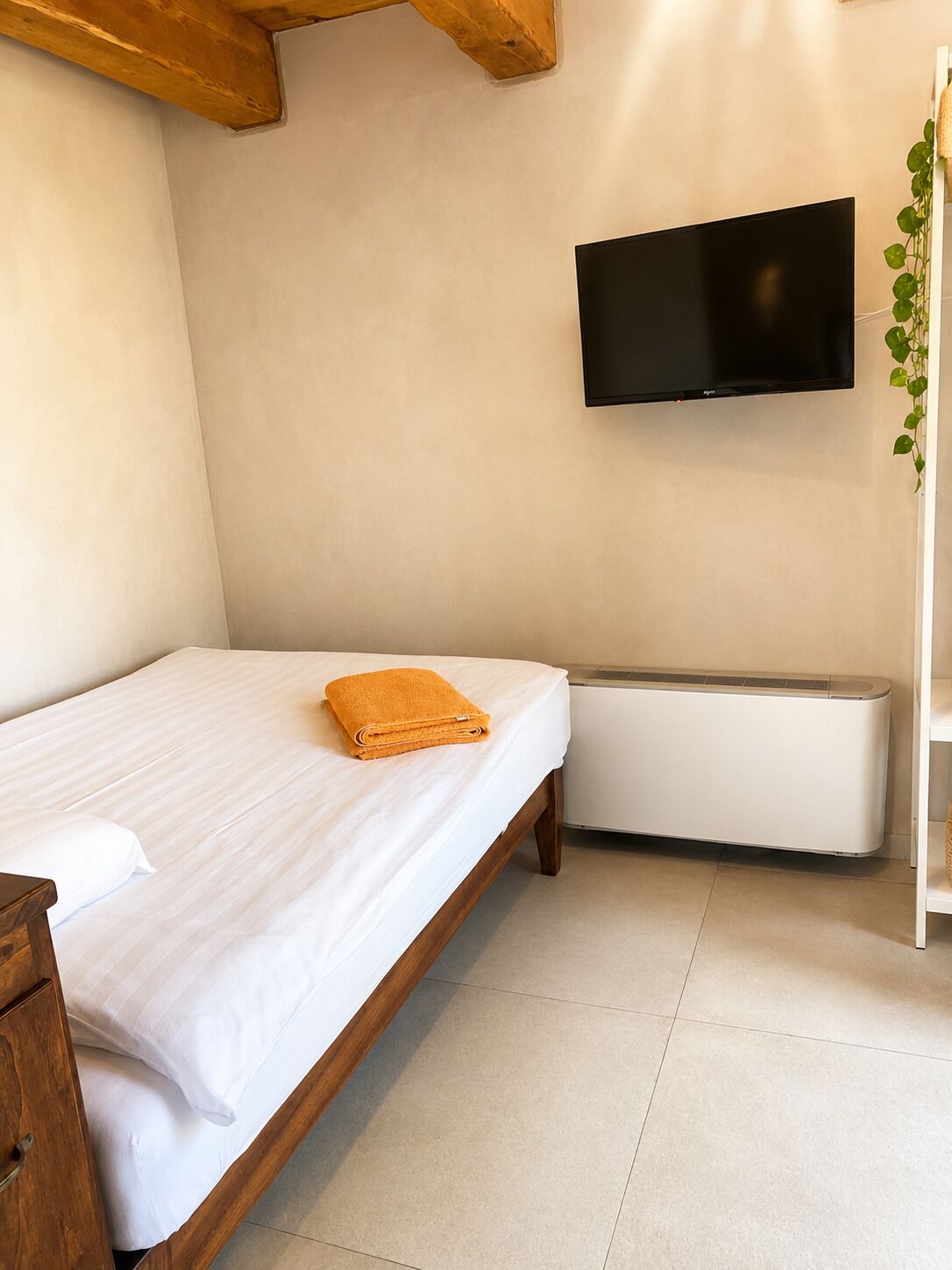 A bedroom with a bed, a TV mounted on the wall, a radiator, and a plant.