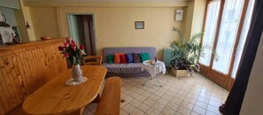 A living room with a wooden dining table, a grey sofa with colorful cushions, a small round table with a white tablecloth, and a potted plant. - Holiday Home "Gîte 1265 in the Lake Region" with Shared Garden and Wi-Fi (Bonlieu)