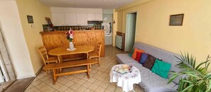 A living room with a wooden dining table, a sofa, and a small round table with a basket and books. - Holiday Home "Gîte 1265 in the Lake Region" with Shared Garden and Wi-Fi (Bonlieu)