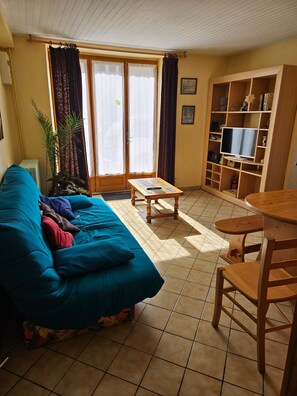 A living room with a sofa, a coffee table, a TV stand, and a dining table with chairs. - Holiday Home "Gîte 1265 in the Lake Region" with Shared Garden and Wi-Fi (Bonlieu)