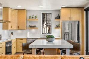 A modern kitchen with wooden cabinets, a stainless steel refrigerator, a marble island, and a dining area with leather chairs. - The Nest | Exclusive Tamarack Perks (Tamarack)