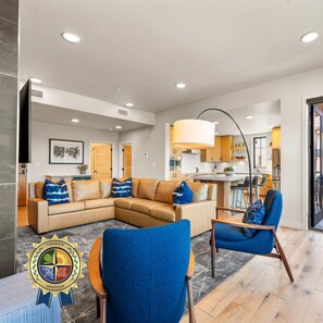 A modern living room with a sectional sofa, a blue armchair, and a dining area with a kitchen in the background. - The Nest | Exclusive Tamarack Perks (Tamarack)