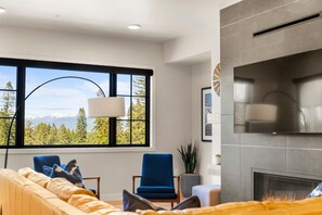 Ein modernes Wohnzimmer mit Kamin, großem Fenster und Blick auf Bäume und Berge, sowie einer Couch mit Kissen.