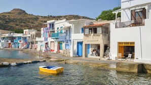 A coastal town with buildings along the water, a small boat, and a hill in the background. - Rota Klima (Milos)