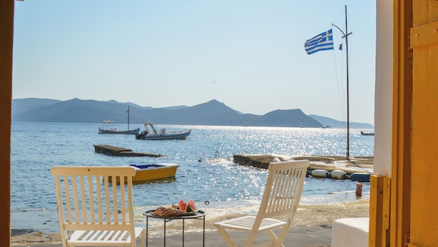 A view from a room with a wooden balcony overlooking a body of water with boats, a flag, and distant mountains. - Rota Klima (Milos)
