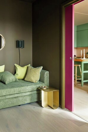 Ein Zimmer mit einem grünen Sofa, einem kleinen goldenen Beistelltisch, einer Wandleuchte und einer Tür zu einem Küchenbereich.