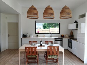 Eine moderne Küche mit weißen Schränken, einem Esstisch mit Stühlen, einem Fenster mit Blick und drei Weidenkorb-Pendelleuchten.