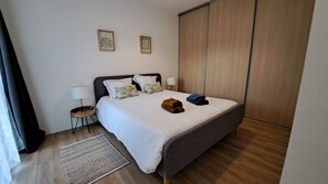 Un lit bien fait, avec des oreillers, une lampe de chevet, une petite table ronde et un tapis tissé. Deux œuvres d’art encadrées ornent le mur.