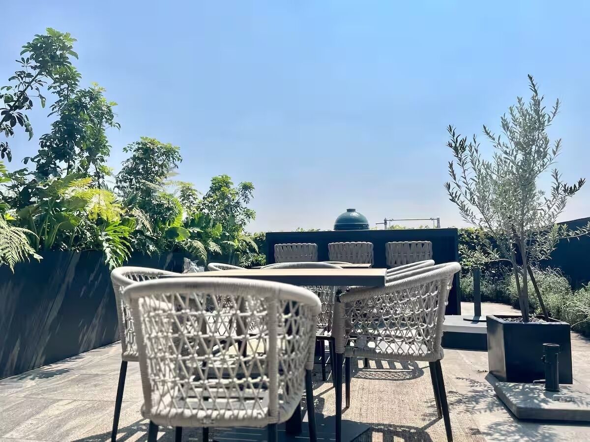 A rooftop patio with wicker chairs and a table, surrounded by greenery and a clear sky.
