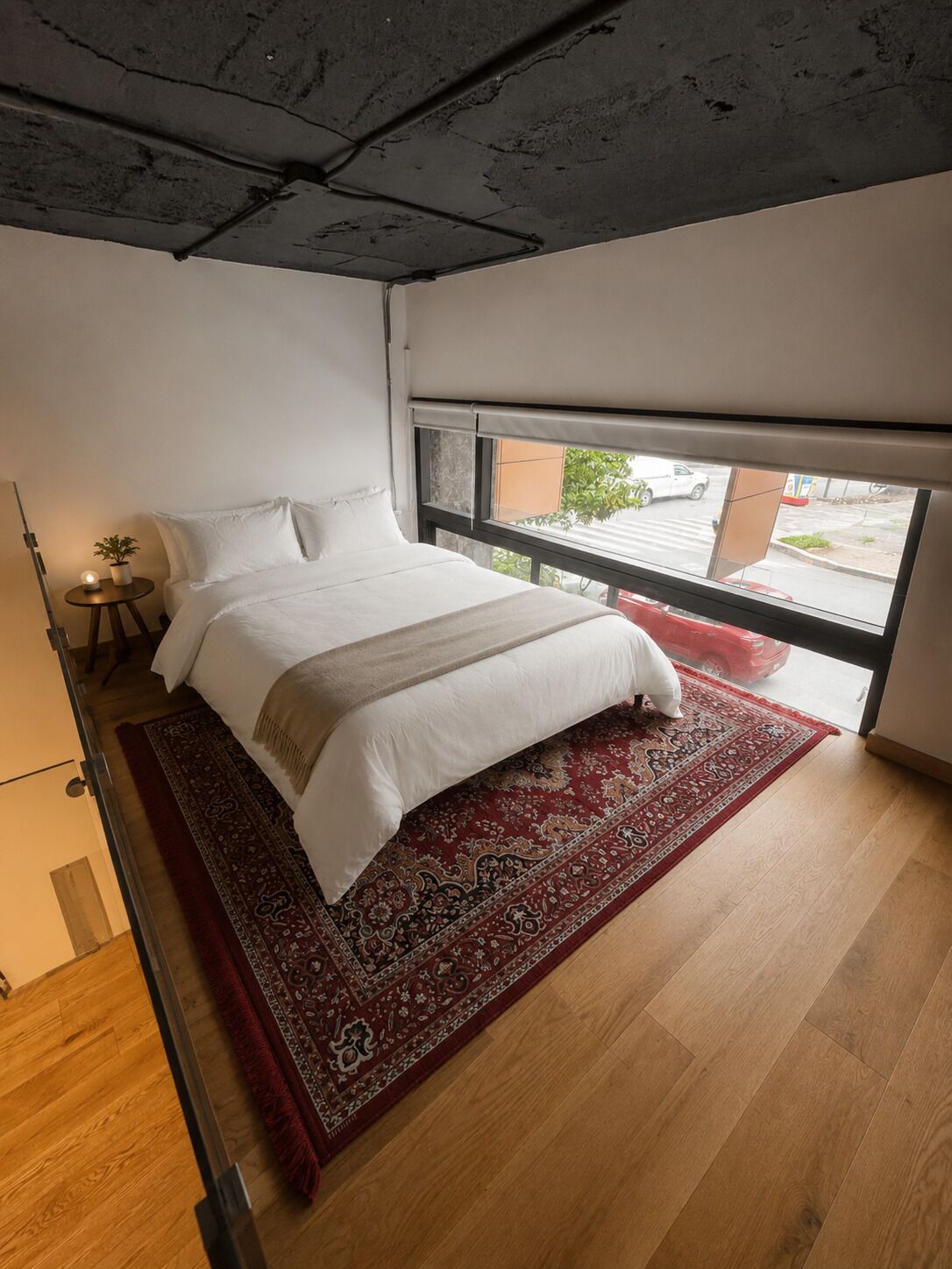 Une chambre à coucher avec un lit, une fenêtre, un tapis rouge et une petite table avec une lampe.