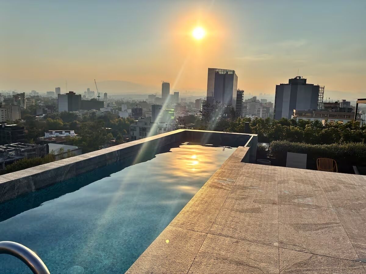 Une piscine à débordement offrant une vue sur le paysage urbain, un ciel dégagé et un soleil couchant à l’horizon.