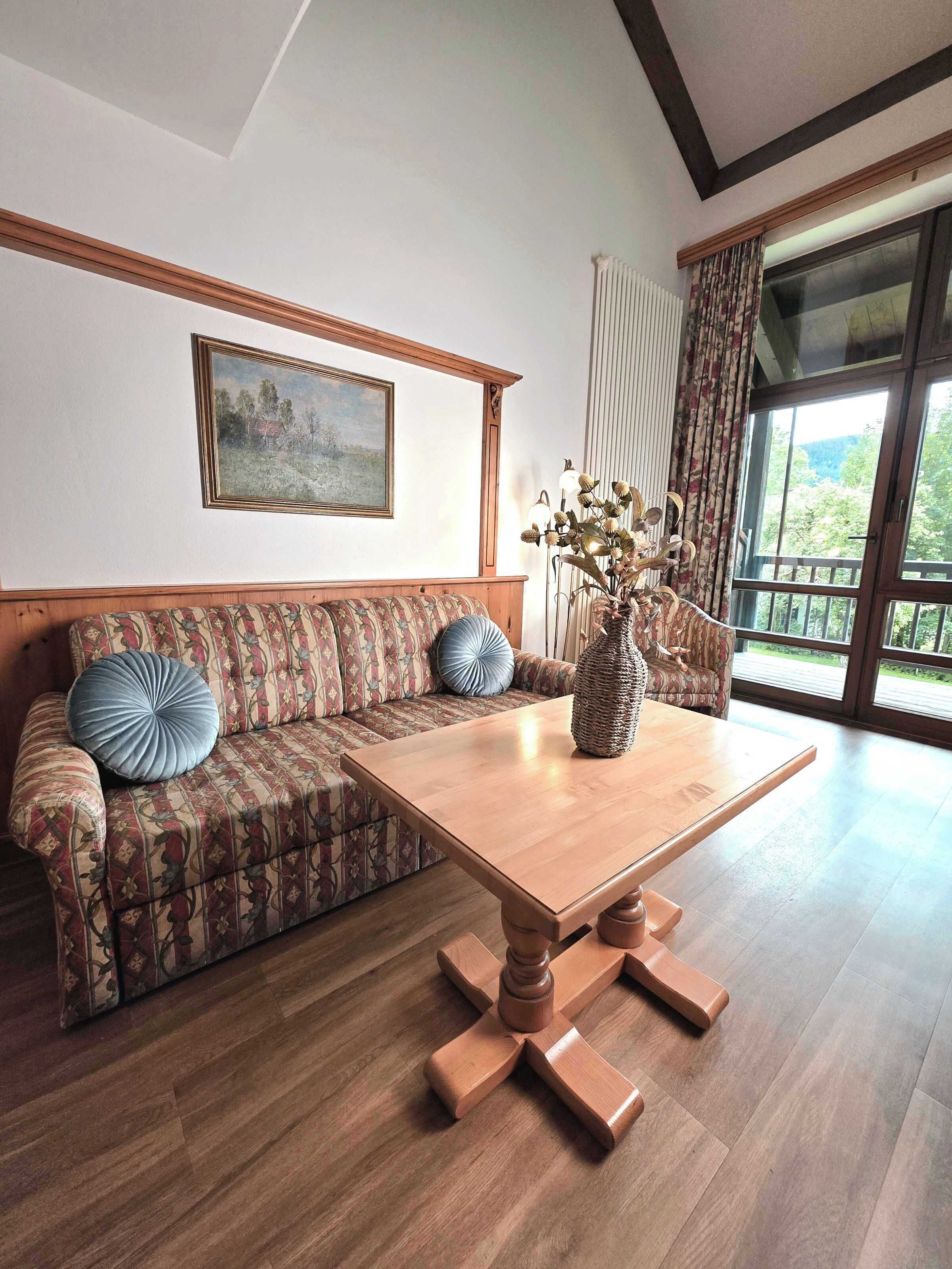 Ein Zimmer mit Holzboden, einer Couch mit gemusterten Kissen, einem Holztisch und einem Bild an der Wand. Ein Fenster mit Vorhängen.