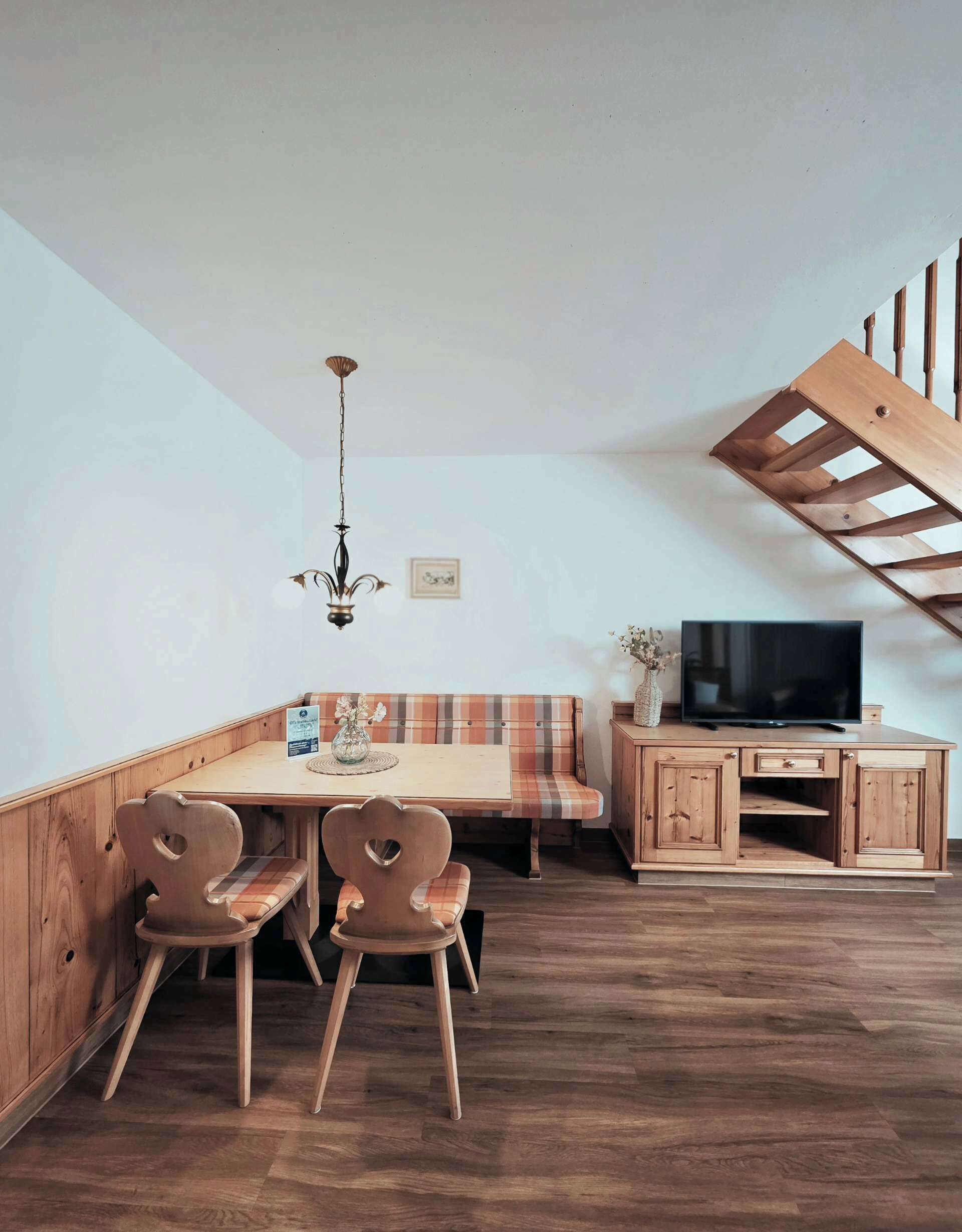A dining area with a wooden table, chairs, and a bench. There is a television on a wooden cabinet and a chandelier hanging from the ceiling.