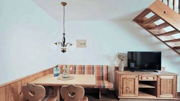 A dining area with a wooden table, chairs, and a bench. There is a television on a wooden cabinet and a chandelier hanging from the ceiling.