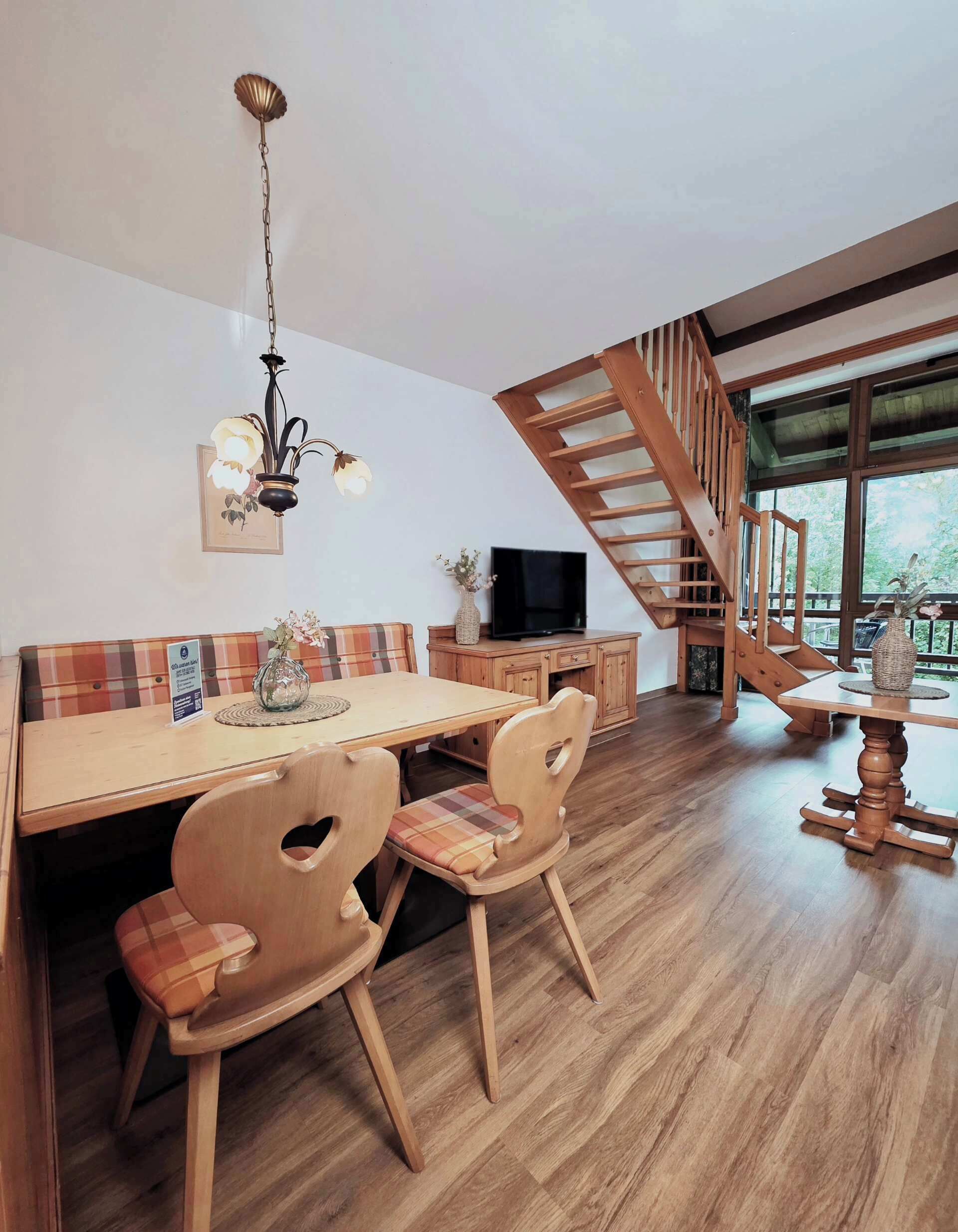 A dining area with a wooden table, chairs, a chandelier, a television, and a staircase.